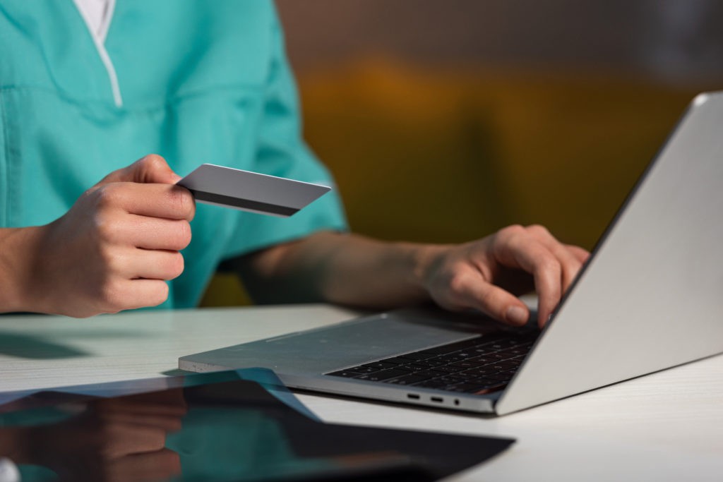 Eine Person in einem grünen OP-Kittel, die vielleicht ihr *Konto für Ärzte* verwaltet, hält eine Kreditkarte in der Hand und tippt auf einem Laptop. Die Szene lässt auf Online-Shopping oder elektronisches Bezahlen schließen. Ein Tablet liegt auf dem Tisch daneben.