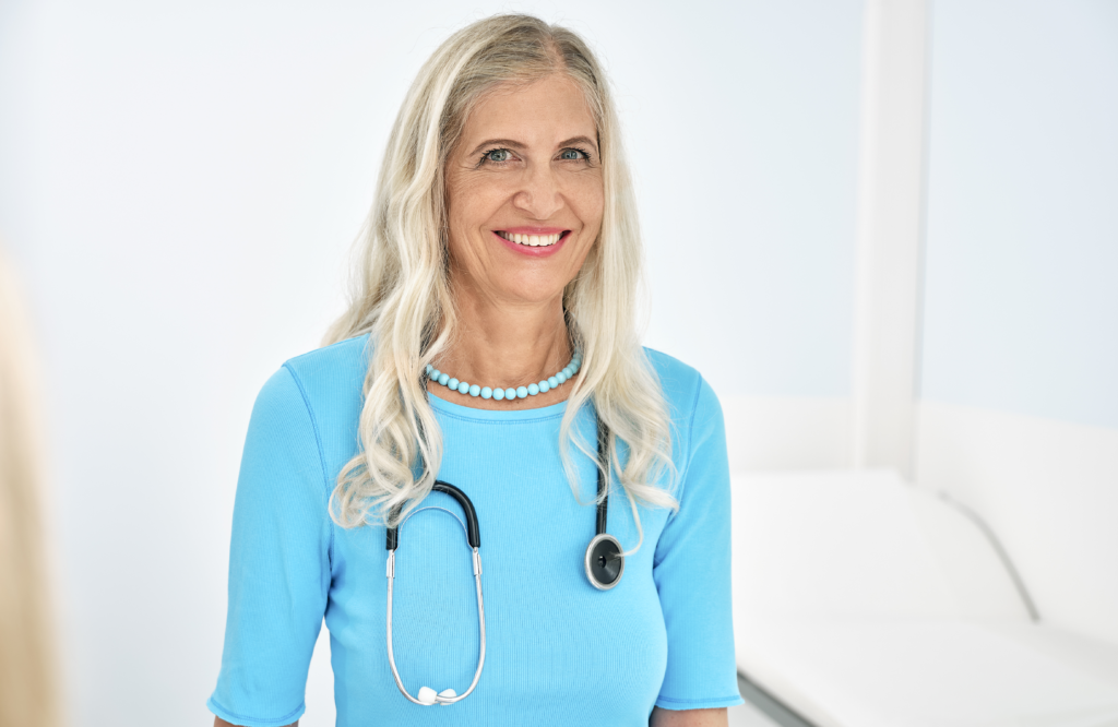 Eine lächelnde Frau mit langen blonden Haaren, einem blauen Hemd und einem Stethoskop um den Hals verkörpert das Bild eines praktizierenden Arztes. Sie steht in einer hellen, sauberen Arztpraxis und strahlt Professionalität und Wärme aus.