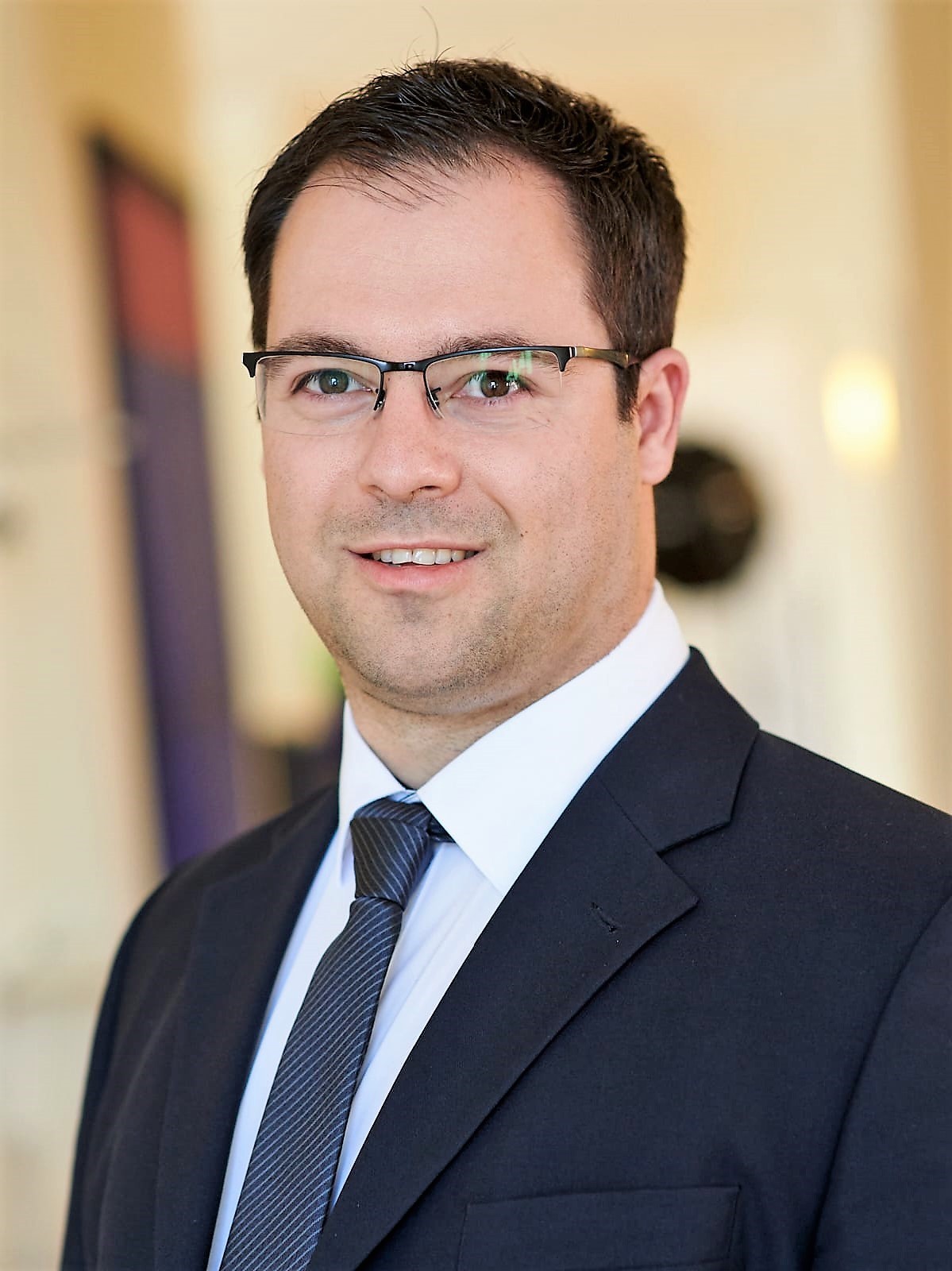 Ein Mann mit Brille und dunklem Anzug, weißem Hemd und gestreifter Krawatte lächelt leicht und denkt vielleicht über seine kürzlich geleistete Steuernachzahlung nach. Der Hintergrund ist leicht verschwommen, und warmes Licht verstärkt die nachdenkliche Stimmung.