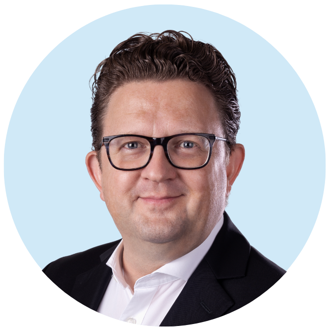 Ein Mann mit kurzen, welligen Haaren und Brille trägt einen dunklen Anzug und ein weißes Hemd. Er posiert vor einem hellblauen Hintergrund und blickt mit neutralem Gesichtsausdruck direkt in die Kamera.