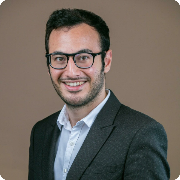 Eine Person mit kurzen dunklen Haaren und Brille lächelt auf einem Porträtfoto. Sie trägt einen dunklen Blazer über einem weißen Hemd mit Kragen vor einem schlichten Hintergrund.