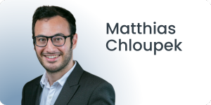 Ein Mann mit Brille, dunklem Haar und Bart lächelt in die Kamera. Er trägt einen dunklen Anzug und ein weißes Hemd. Der Hintergrund ist ein Farbverlauf von Blau nach Weiß, neben ihm ist der Text Matthias Chloupek zu sehen.