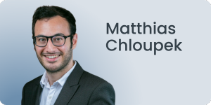 Eine lächelnde Person mit kurzen dunklen Haaren und Brille, die einen dunklen Anzug und ein weißes Hemd trägt. Der Hintergrund ist ein Farbverlauf von Blau nach Grau mit dem Namen Matthias Chloupek darauf geschrieben.