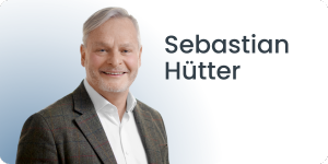 Ein lächelnder Mann mit grauem Haar, der ein weißes Hemd und einen karierten Blazer trägt, steht vor einem Hintergrund mit Farbverlauf und dem Text „Sebastian Hütter“ neben ihm.