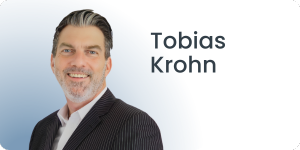 Ein Mann mit kurzen Haaren und Bart lächelt und trägt einen dunklen Nadelstreifenanzug mit weißem Hemd. Der Hintergrund ist ein Farbverlauf aus Blau und Weiß. Der Text rechts lautet Tobias Krohn.